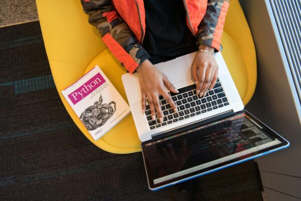 A developer typing code on a laptop with a Python book beside in an office.